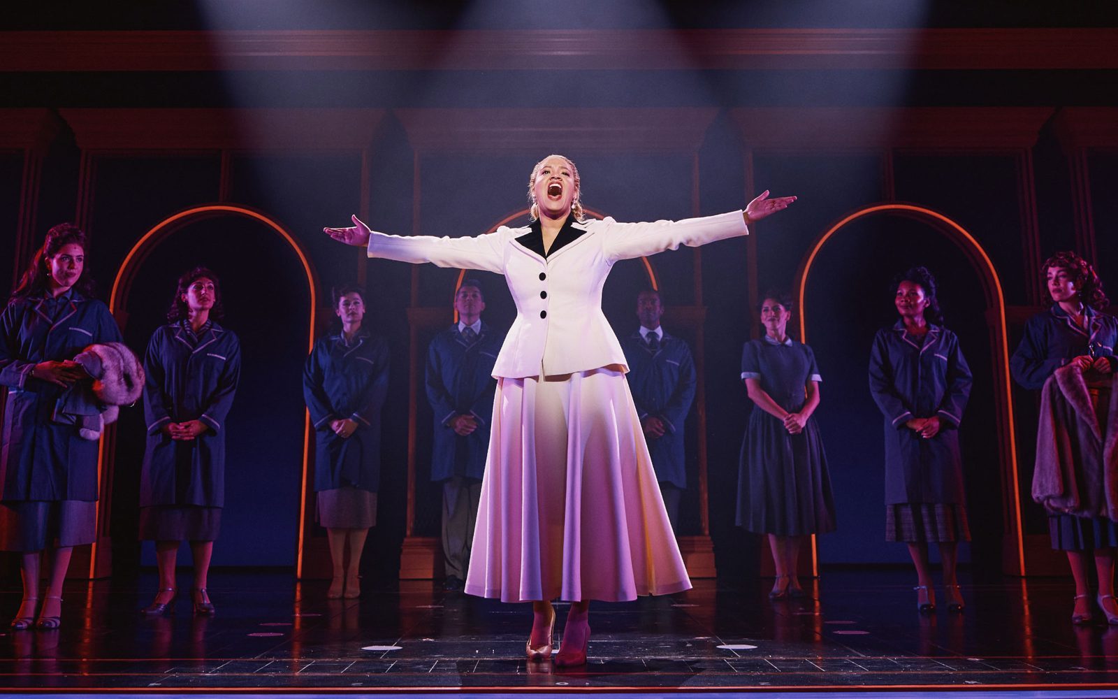 Shereen Pimentel as Eva standing in the spotlight with her arms outstretched while cast members look on.