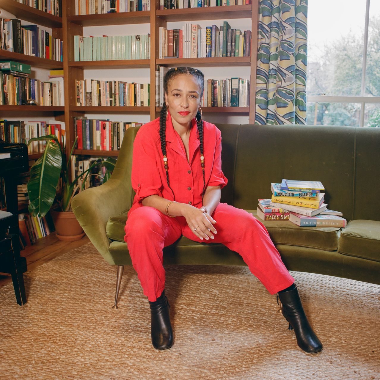 Zadie Smith in a red jumpsuit seated on a muted green couch in front of a bookshelf in her living room.
