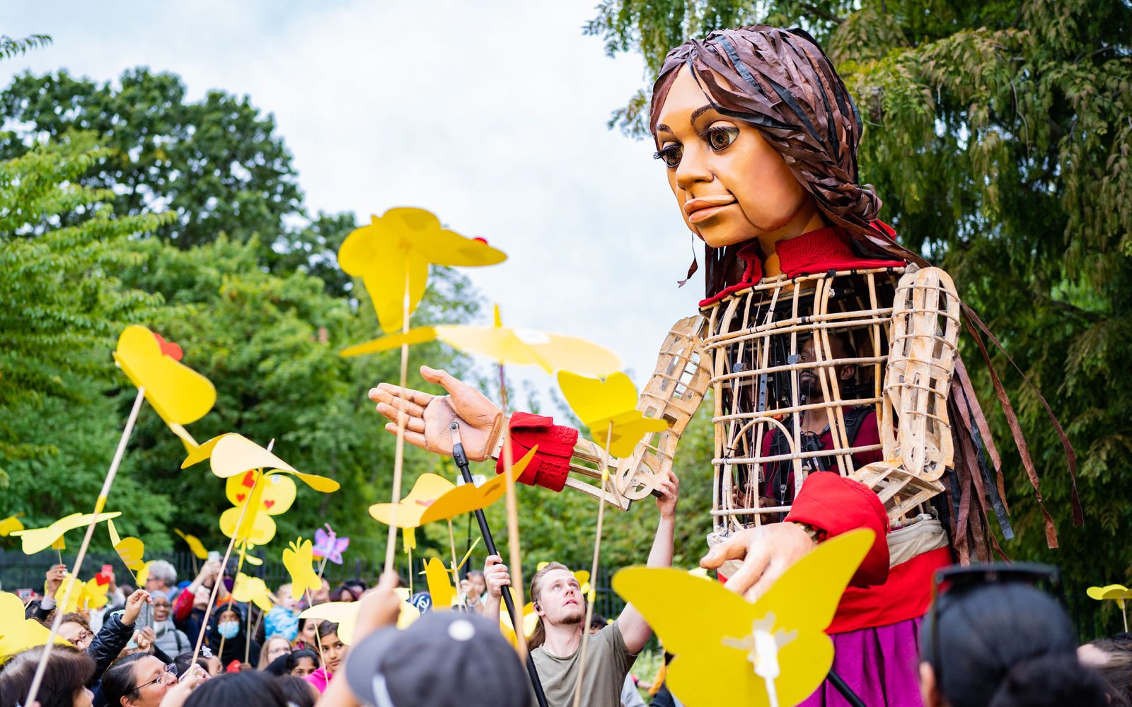 Little Amal in a clearing between trees as people raise yellow butterflies to her.