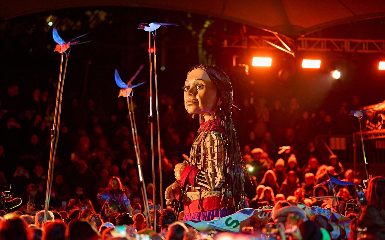 Little Amal looks over a crowd to three bird puppets flying towards her.