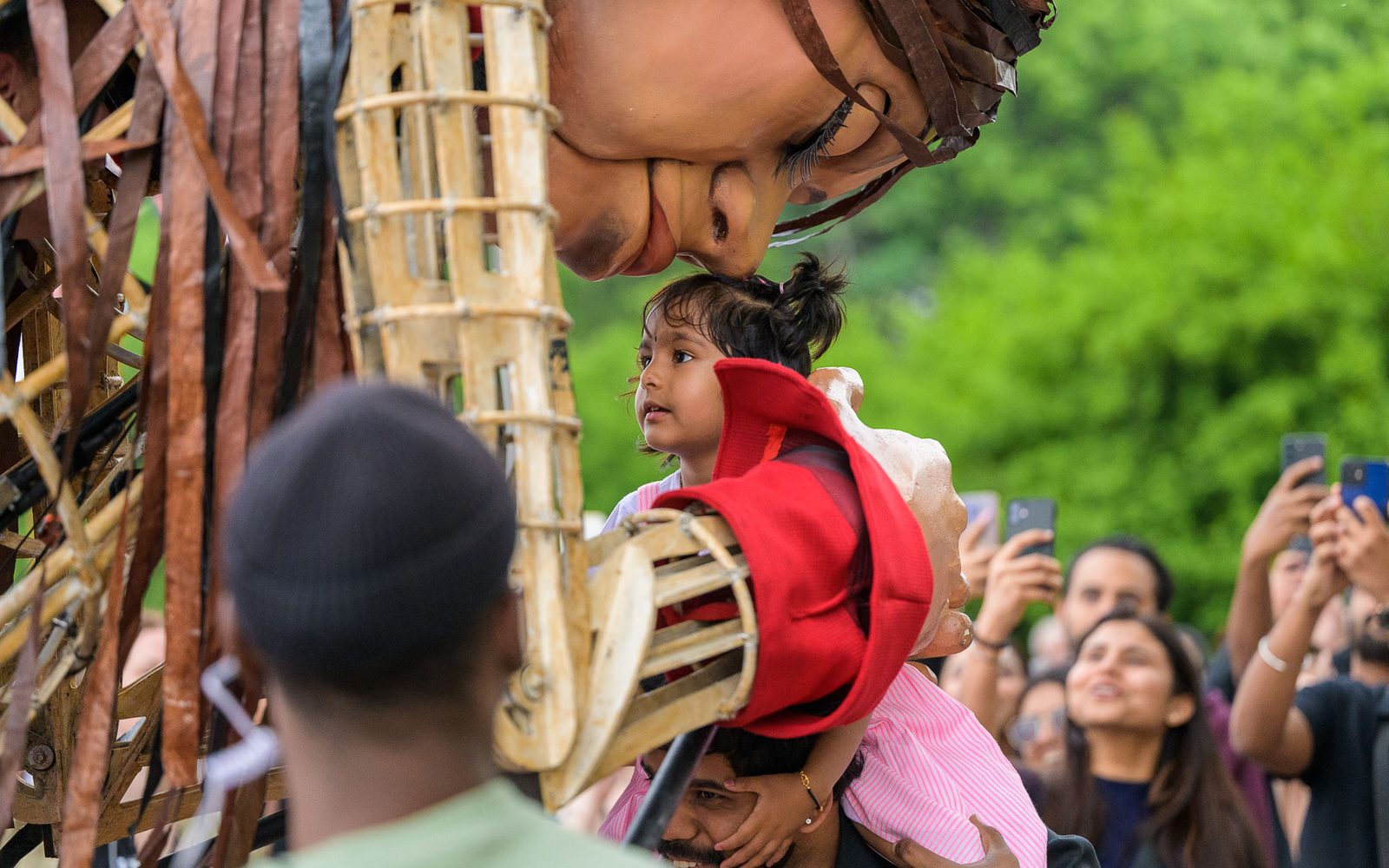 Little Amal bends over to put her head to a child’s.