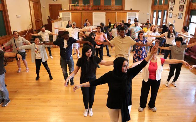 A group of people stand staggered in rows and raise their arms by their sides.