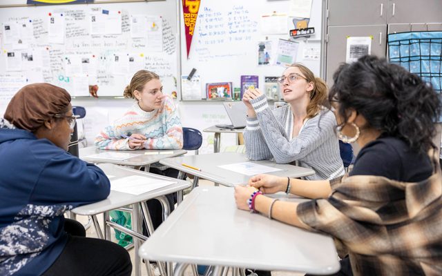 Four students sit in a group at desks in a brightly lit classroom. One of them looks upward as she speaks while the other three watch.
