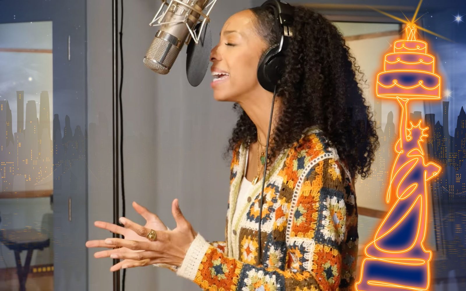 Christiani Pitts in a patterned knit cardigan raises her hands as she sings into a studio microphone.