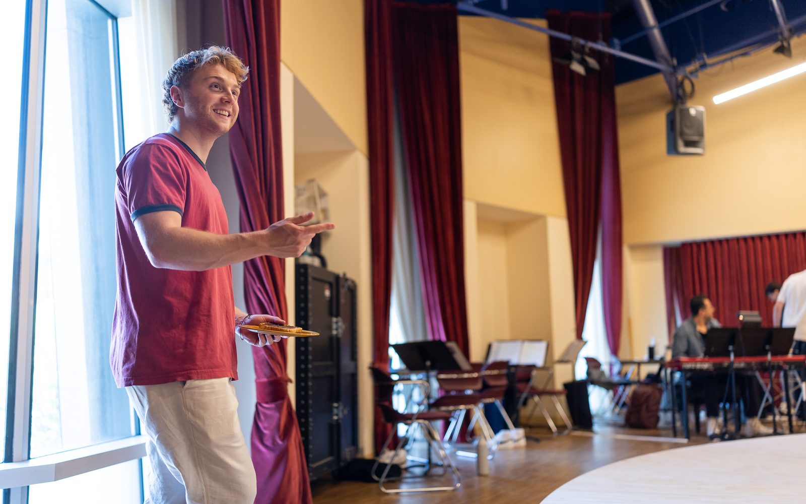 Sam Tutty in a red T-shirt and white pants holds a plate with a slice of toast on it while smiling to someone off camera.