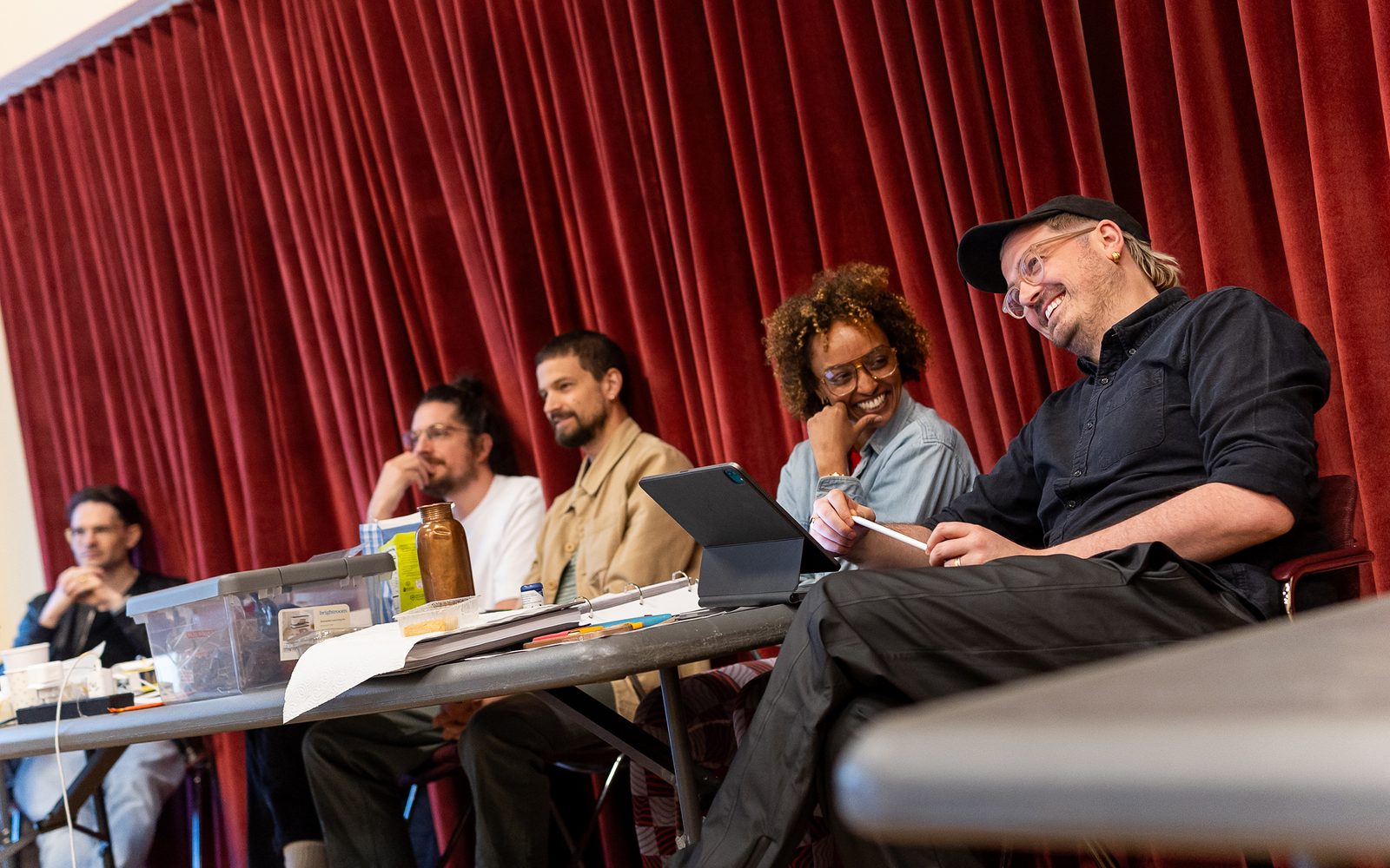 Sitting at a folding table in front of a red curtain, Jim Barne and Kit Buchan watch action in the rehearsal room, while Asmeret Ghebremichael and Tim Jackson smile at each other.