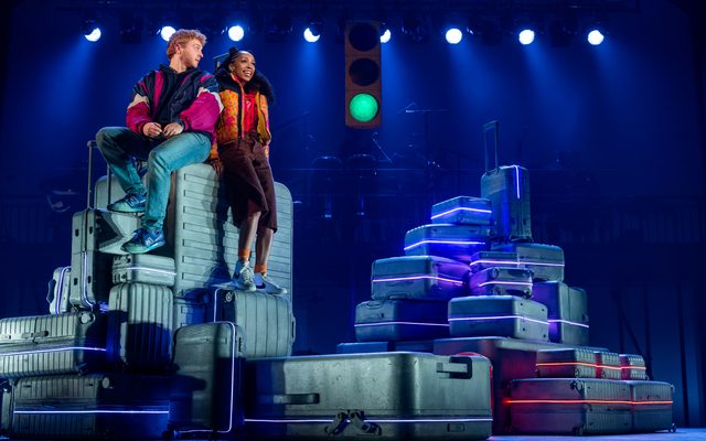 Sam Tutty in a magenta windbreaker and Christiani Pitts in an orange-and-magenta puffer jacket sit on top of a stylized stack of suitcases.