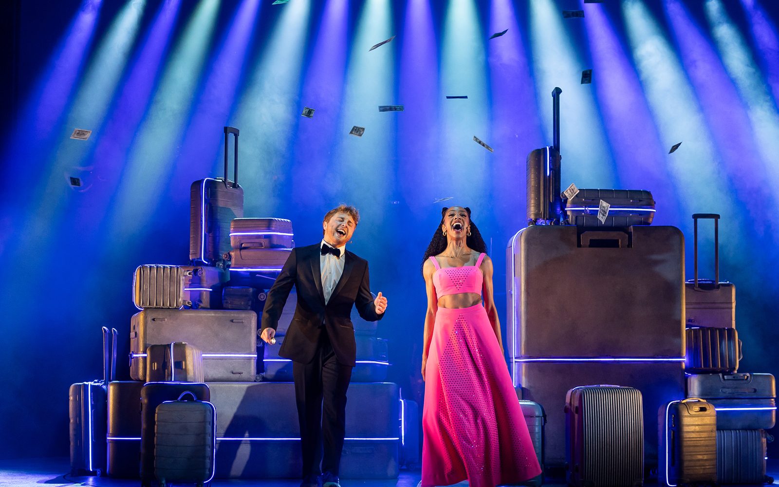 Sam Tutty in a tuxedo and Christiani Pitts in a pink gown adorned with rhinestones stand center stage singing loudly under a wash of blue and white lights and dollar bills falling down on them.