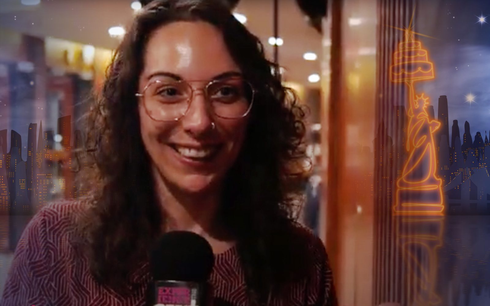 A woman with dark wavy hair and glasses speaks into a microphone.