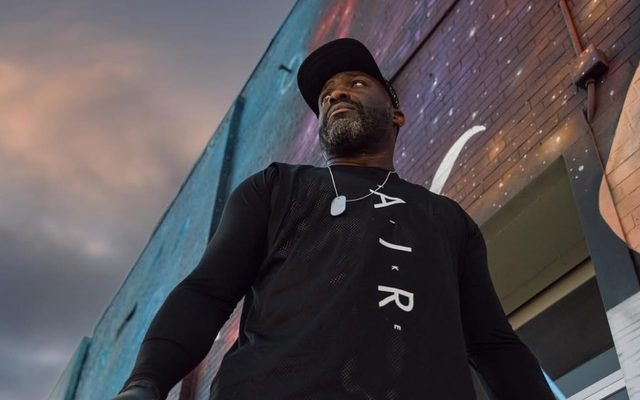 Problak in a black shirt and cap seen from below standing in front of a star-studded mural.