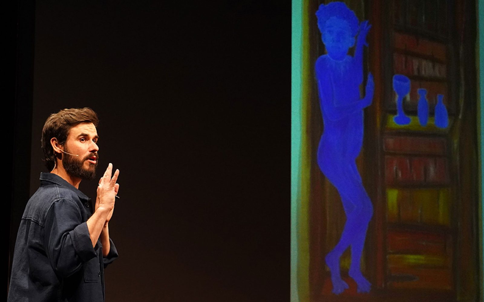 Sam Kissajukian in a dark grey flannel shirt raises his arms while speaking. To his right is a large paintings that features a bright blue human figure next to a black and green bookshelf.