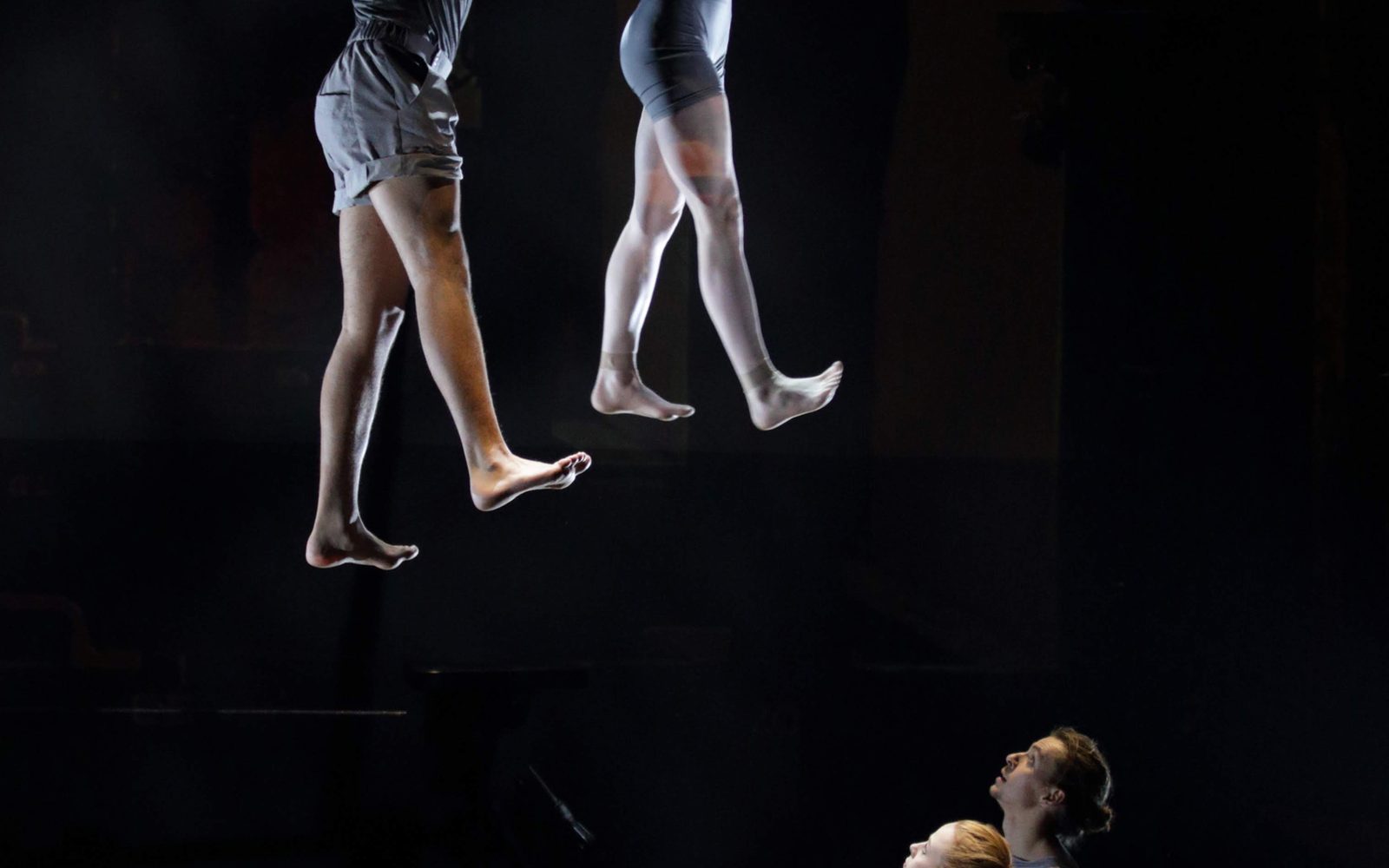 Two acrobats hang side-by-side from a trapeze by their hands with their feet flattened as if they're walking. Two performers on the ground look up at them.